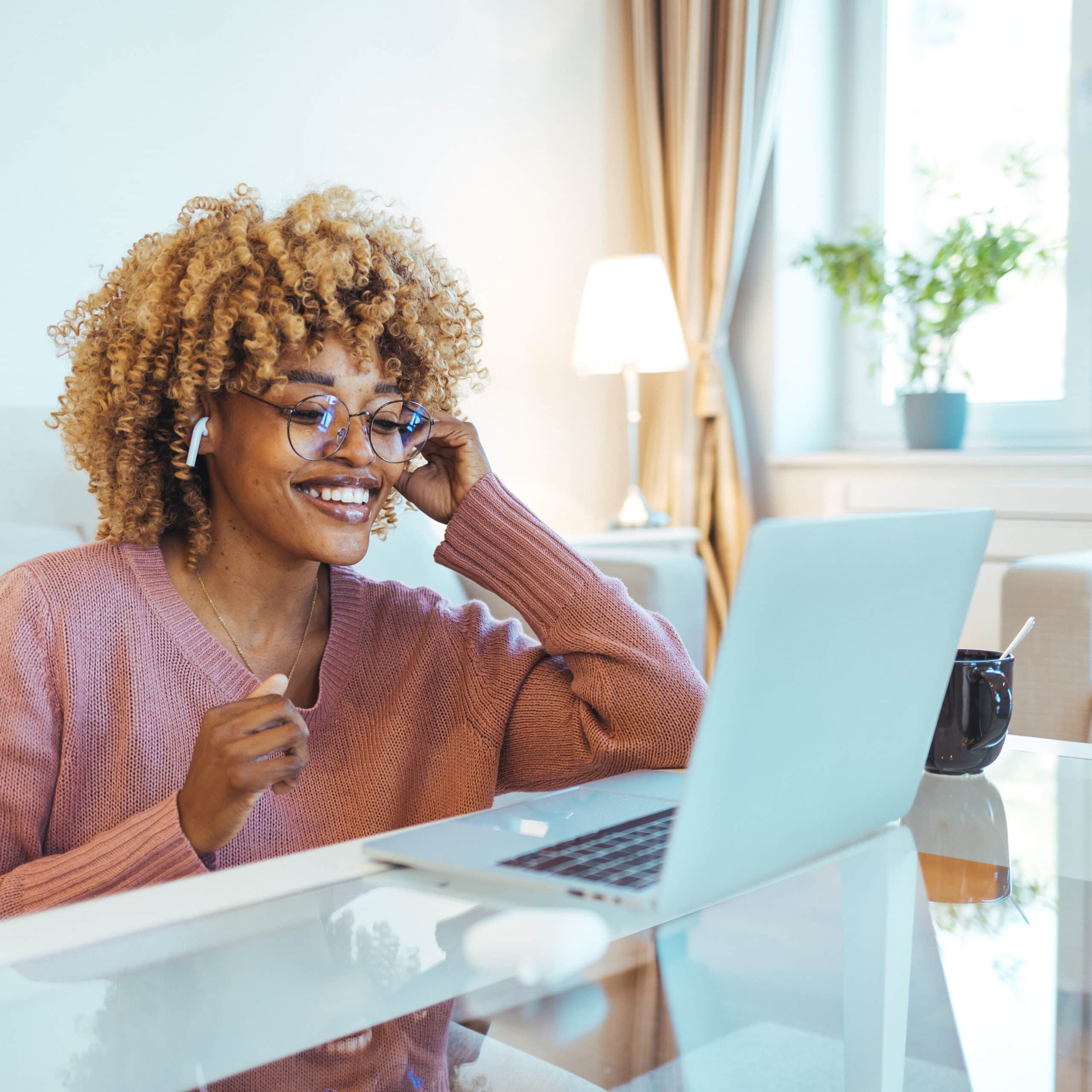 Smiling African American Woman Wearing Glasses and Wireless Earphones Makes a Video Call on her Laptop Computer at her Home Office. Smiling businesswoman in a video call using laptop in the office